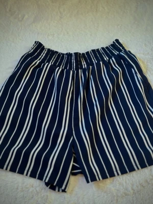 Randiga blå shorts med resårmidja - Snygga blå shorts med vita vertikala ränder och bekväm resår i midjan. Perfekta för varma dagar och enkel att matcha med en t-shirt eller linne. Luftigt material som känns skönt mot huden.