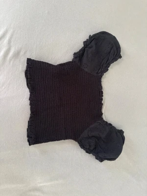 Svart blus med puffärmar - Trendig svart blus med smockad kropp och korta puffärmar i broderad bomull. Perfekt att styla med jeans eller kjol🤍