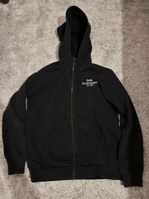 Svart Peak Performance hoodie - Svart hoodie med dragkedja från Peak Performance. Klassisk design med broderad logga och text 'Est. 1986' på bröstet. Utrustad med huva och kängurufickor framtill. Perfekt för en avslappnad och sportig stil.
