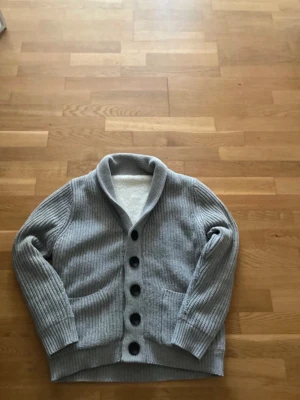 Grå ribbstickad kofta med stora knappar - Snygg grå ribbstickad kofta med stora svarta knappar och två fickor framtill. Koftan har en bred krage och långa ärmar med ribbade muddar. Perfekt för lager-på-lager och chill dagar.