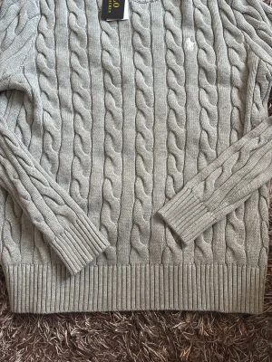 Ljusgrå kabelstickad tröja Gant - Kabelstickad tröja i ljusgrått från Gant med rund halsringning och ribbade muddar vid ärmslut och nederkant. Klassisk design med diskret logga broderad på bröstet. Perfekt för en stilren och avslappnad look.