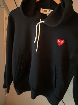 Svart hoodie från Comme des Garçons Play - Svart hoodie med huva och vita dragsnören från Comme des Garçons Play. På bröstet sitter det ikoniska röda hjärtat med ögon. Hoodien har känguruficka och är tillverkad i mjukt material, perfekt för en avslappnad streetstyle. Ganska liten i storlek 