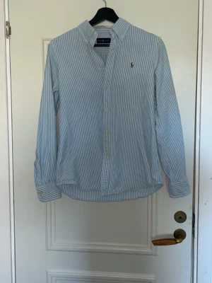 Blårandig skjorta från Ralph Lauren - Klassisk blårandig skjorta från Ralph Lauren med button-down krage och broderad logga på bröstet. Skjortan  långa ärmar, vita knappar och är tillverkad i 100%  mjuk bomull. Perfekt inför sommaren🙌🏻 Skick 10/10