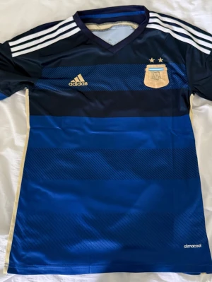 Argentina Adidas fotbollströja blå - Snygg blå fotbollströja från Adidas med Argentinas landslagsemblem och två stjärnor på bröstet. Tröjan har vita ränder på axlarna, guldiga detaljer och är tillverkad i lätt och ventilerande climacool-material. Perfekt för fotbollsträning eller match.