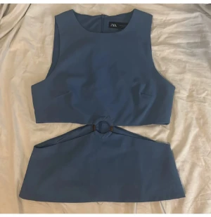 Blå topp från Zara - Säljer en blå croppad topp från Zara med ärmlös design och cool cut-out detalj i midjan med ring. Perfekt för dig som vill ha en trendig och unik look. Materialet känns mjukt och följsamt mot kroppen.