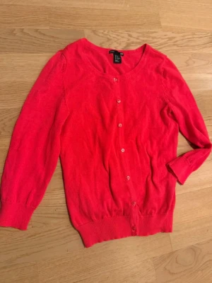 Röd kofta från H&M med knappar - Snygg röd kofta från H&M med rund halsringning och knäppning framtill. Koftan har långa ärmar och ribbade muddar vid ärmslut och nederkant. Perfekt att slänga över en t-shirt eller klänning.