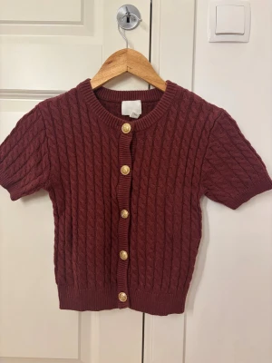 Kortärmad vinröd kabelstickad kofta - Kortärmad kabelstickad kofta i vinrött med rund hals och dekorativa guldknappar framtill. Ribbstickade muddar vid ärmslut och nederkant. Perfekt att styla över en topp eller klänning för en trendig look. Pris kan diskuteras
