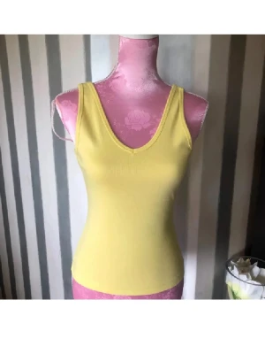 Gul ribbad linnetopp S - Snygg gul linnetopp med v-ringning både fram och bak. Toppen är ribbad och har en tight passform, perfekt för att styla med jeans eller kjol. Tillverkad i mjuk bomull som känns skön mot huden.