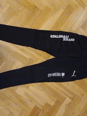 Svarta Puma mjukisbyxor med tryck - Svarta mjukisbyxor från Puma med vita tryck på benen, bland annat texten 'OFFICE IF-FRÅNTER' och 'MI FEEDBACK'. Byxorna har smal passform och resår vid bensluten. Perfekta för en avslappnad och sportig stil. Skriv för fler bilder.