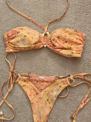 Somrig bandeau bikini sett med knyt från Lounge - Hej!🌸Säljer en snygg bandeau-bikini med knytning både på toppen och trosan. Bikinin har ett färgglatt mönster i gula, orange och beige toner med blommiga detaljer. Materialet är mjukt och stretchigt, perfekt för soliga sommardagar på stranden. Det finns lite slitage på två små spännen (syns på bilden), men det är knappt märkbart. Bikinin är sparsamt använd! Nypris för trosan är 499 kr och för överdelen 525 kr.🩷