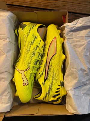 Gula Puma fotbollsskor med svart mönster - Säljer ett par helt nya, riktigt snygga fotbollsskor från Puma i neon-gult med svarta och röda detaljer. Skorna har ett sportigt mönster, klassisk snörning och dobbar för gräsplan. Perfekta för dig som vill sticka ut på planen och få bra grepp vid snabba vändningar.