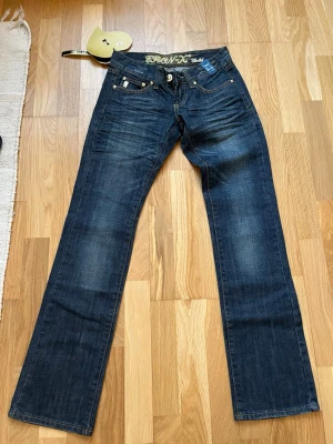 Mörkblå bootcut jeans från Cron-X - Snygga mörkblå bootcut jeans från cron-x med guldfärgade detaljer på bakfickorna. Jeansen har låg midja och lätt slitna partier framtill för en cool look. Oanvända och lappen sitter kvar. W25 och L32