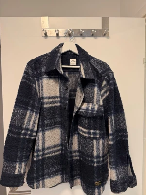 Rutig overshirt från Zara i blått - Säljer en snygg och trendig overshirt från Zara i relaxed fit. Den har ett rutigt mönster i mörkblått, vitt och grått, samt en bröstficka och klassisk krage. Materialet är mjukt och lite fluffigt, perfekt för lager-på-lager-stil.