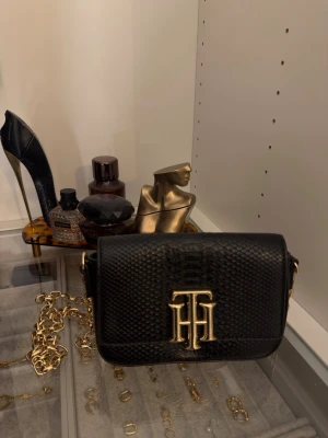 Svart handväska från Tommy Hilfiger - Snygg svart handväska i läder med reptilmönster och guldfärgade detaljer. Väskan har en guldig kedja som axelband och ett stort TH-logo i guld på framsidan. 