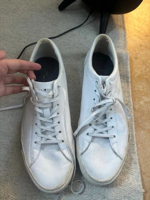 Vita Tommy Hilfiger sneakers - Snygga vita sneakers från Tommy Hilfiger med klassisk låg profil och lädermaterial. Skorna har vita snören, diskret logotyp på sidan och en stilren design med små färgdetaljer på sulan. Perfekta för dig som gillar en clean och modern look. Endast använda en kväll 