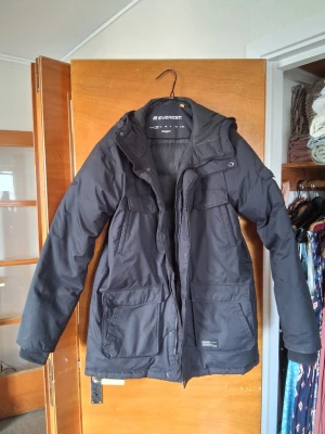 Svart parkasjacka från Everest - Säljer en svart parkasjacka från Everest med huva och flera praktiska fickor framtill. Jackan har dragkedja och knappar, samt ett slitstarkt yttertyg som står emot kyla och vind. Perfekt för kalla dagar när du vill hålla dig varm och bekväm.