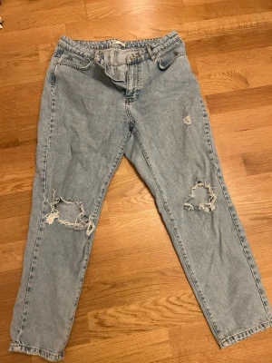 Ljusblå  jeans - Säljer ett par ljusblå jeans med slitna detaljer och hål på båda knäna. Jeansen har hög midja. Perfekta för en avslappnad och trendig look.