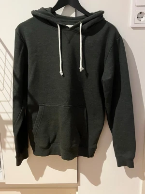 Mörkgrön hoodie från Pier One - Mörkgrön hoodie från Pier One med klassisk känguruficka och vita dragsnören i huvan. Mjuk bomullsblandning och ribbade muddar vid ärmslut och nederkant. Perfekt för en chill och avslappnad stil.