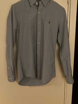 Ljusblå skjorta från Ralph Lauren - Klassisk ljusblå skjorta från Ralph Lauren med button-down krage och broderad logga på bröstet. Skjortan har långa ärmar, knappar framtill och är tillverkad i mjuk bomull. Perfekt för en clean och stilren look.