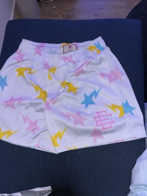 Vita Eric Emanuel shorts med stjärnor och blixtar - Säljer ett par vita shorts från Eric Emanuel med färgglada stjärnor och blixtar i blått, rosa och gult. Shortsen har elastisk midja och EE-logga broderad framtill. Perfekta för en färgstark och sportig look.