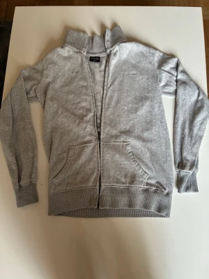 Ljusgrå zip-hoodie från On The Peak - Snygg ljusgrå hoodie från On The Peak med dragkedja framtill och två stora fickor. Tröjan har ribbade muddar och krage, samt diskret logga på bröstet. Perfekt för en chill och avslappnad stil.