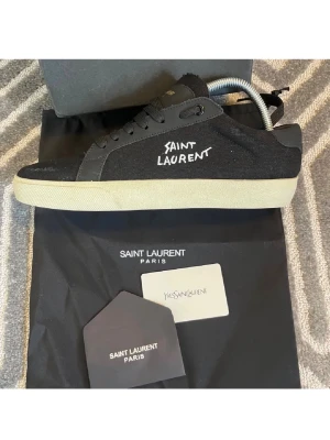 Svarta Saint Laurent sneakers - Svarta sneakers från Saint Laurent med vit broderad logga på sidan och guldfärgad text på (tungan) . Skorna har en klassisk låg siluett, svarta skosnören och vit sula. Detaljer i mocka. Perfekta för dig som gillar stilrena och exklusiva sneakers. Bara använda ett få gånger. Box och dustbag hänger med på köpet. Bara höra av dig om du har några frågor!