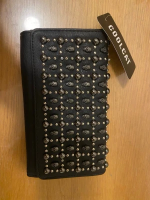 Svart plånbok med nitar från Coolcat - Snygg svart plånbok från Coolcat i konstläder med silverfärgade nitar och små dödskalle-detaljer på framsidan. Plånboken har flera fack för kort, sedlar och mynt, samt dragkedja inuti. Perfekt accessoar för dig som gillar en edgy stil.