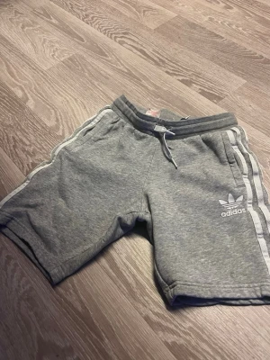Grå adidas shorts med snörning - Säljer ett par grå adidas shorts med klassiska vita ränder längs sidorna och adidas-logga framtill. Shortsen har snörning i midjan och är tillverkade i mjuk bomull, perfekta för en avslappnad stil.