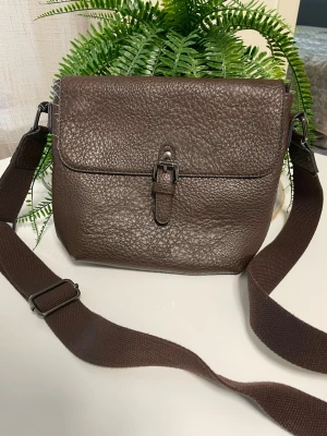 Brun axelväska i läderlook - Snygg brun axelväska i kraftigt, strukturerat konstläder med justerbar textilrem. Väskan har ett lock med spänne framtill och en praktisk dragkedjeficka på baksidan. Perfekt för dig som vill ha en stilren och funktionell väska till vardags.