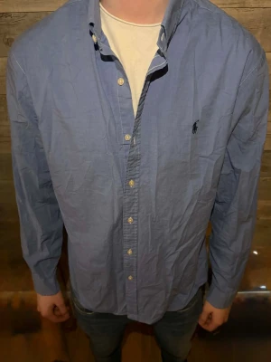 Blå skjorta från Polo Ralph Lauren - Klassisk blå skjorta från Polo Ralph Lauren med button-down krage och broderad logga på bröstet. Skjortan har långa ärmar, knäppning framtill och är tillverkad i bomull. Perfekt för en stilren look.