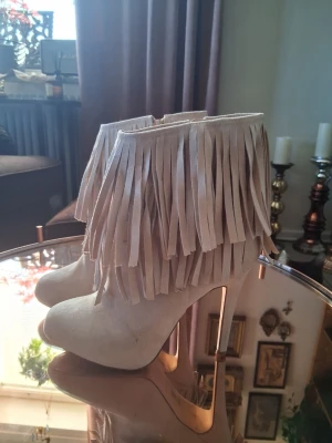 Beiga stövletter med fransar och klack - Snygga beiga stövletter med höga klackar och coola fransdetaljer runt skaftet. Tillverkade i mockaimitation som ger en mjuk känsla och trendig look. Perfekta för dig som vill sticka ut med din stil och gillar boho-vibbar. 😃 Endast använda 1 gång.