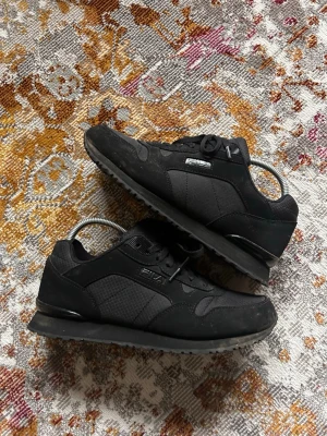 Svarta Fila sneakers i klassisk stil - Svarta sneakers från Fila med lågt skaft, snygga detaljer i textil och mockaimitation. Skorna har en sportig siluett, svarta snören och diskret Fila-logga på sidan. Yttersulan är mönstrad för bra grepp och skon har en stilren, tidlös look.