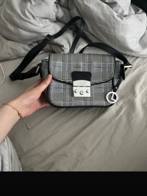 Rutigt axelväska från L.Credi - Snygg axelväska från L.Credi i grått och svart rutigt tyg med svarta detaljer. Väskan har ett silverfärgat lås framtill och en justerbar axelrem i svart. Dekorativt metallhänge med L-logga. Perfekt för att lyfta din outfit. Jag fick den som present och har aldrig använt den. Ute kostar den ca 1000 kr