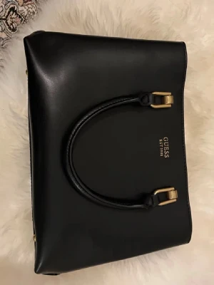 Svart handväska från Guess - Stilren svart handväska från Guess med guldfärgade detaljer och dubbla handtag. Väskan är tillverkad i slätt konstläder och har en praktisk dragkedja på baksidan. Guess-loggan i guld pryder framsidan. Perfekt för dig som gillar klassisk och elegant design.