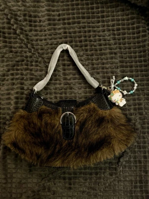 Brun fluffig handväska - Säljer en brun fluffig handväska med svarta detaljer i fejkläder med krokodilmönster. Väskan har ett brett handtag i tyg och ett dekorativt spänne framtill. En söt nyckelring med pärlor och figur hänger på sidan för extra stil.