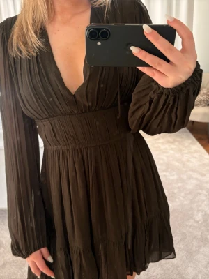 Svart omlottklänning med v-ringning - Säljer en svart kortklänning med v-ringning och långa, transparenta ärmar. Klänningen har omlottdesign, markerad midja och lätt rynkade detaljer. Materialet känns lätt och flowy, perfekt för dig som gillar en chic och stilren look.