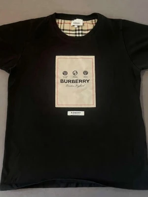 Svart Burberry t-shirt  - Svart t-shirt från Burberry med klassisk logga och tryck framtill. T-shirten har rund hals och korta ärmar. Insidan av kragen har det ikoniska Burberry-rutiga mönstret. Perfekt för dig som gillar stilrena och exklusiva plagg.