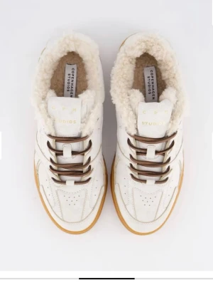 Vita sneakers från Copenhagen Studios - Snygga vita sneakers från Copenhagen Studios med bruna snören, beige foder i fuskpäls och gummisula i ljusbrun färg. Ovandelen är i läder med perforerade detaljer och logga på plösen. Perfekta för dig som vill ha en unik och trendig look. Helt oanvända 