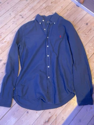 Blå skjorta från Polo Ralph Lauren - Klassisk blå skjorta från Polo Ralph Lauren med knappar framtill och button-down krage. Skjortan har lång ärm och en liten röd broderad logga på bröstet. Perfekt för en stilren look och enkel att matcha med jeans eller chinos.