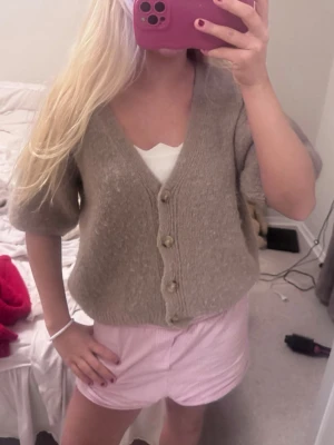 Kortärmad kofta - Mysig beige kofta med korta ärmar och tre knappar framtill. Koftan har en djup v-ringning och är stickad i ett mjukt material som ger en avslappnad look. Perfekt att slänga över ett linne för en chill stil. Från Italien i mohair