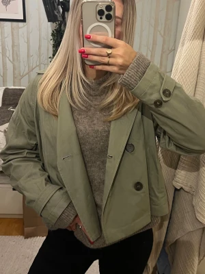 Kort grön trenchcoat - Snygg kort trenchcoat i ljusgrönt med bred krage och knappar. Jackan har en boxig passform och klassiska detaljer som axelklaffar och justerbara ärmslut. Perfekt för dig som vill ha en trendig och stilren look. Passar xs-m❤️