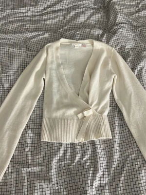 Vit kofta från Gina Tricot - Säljer en krämvit stickad omlottkofta från Gina Tricot med långa ärmar och bred ribbad kant nedtill. Koftan har knytband i midjan och en djup v-ringning. Perfekt att styla över ett linne eller t-shirt för en chill look.