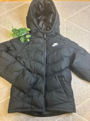 Svart pufferjacka från Nike - Säljer en svart pufferjacka från Nike med huva och dragkedja framtill. Jackan har ett stilrent Nike-logotyptryck på bröstet och är tillverkad i ett glansigt, vadderat material som håller dig varm under kalla dagar. Perfekt för dig som vill ha en sportig och trendig look.