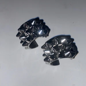 Silvriga Edblad örhängen med nitar - Coola örhängen i silverfärgad metall med ett unikt designmönster av små nitar över hela ytan. De har en halvcirkelformad stil och stängs med klassisk ploppbaksida. Perfekta för dig som gillar edgy accessoarer och vill sticka ut.