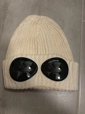 Ljusbeige ribbstickad mössa C.P. Company - Säljer en ljusbeige ribbstickad mössa från C.P. Company med ikoniska svarta glasögonlinser på uppviket. Tillverkad i mjuk ullblandning och har en unik streetwear-look som sticker ut. Perfekt accessoar för dig som vill ha något extra till din outfit.