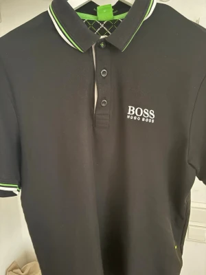 Svart pikétröja från Hugo Boss - Klassisk svart pikétröja från Hugo Boss med vit och grön rand på krage och ärmslut. Tröjan har knappar framtill och logga broderad på bröstet och i nacken. Perfekt till en clean och sportig stil.