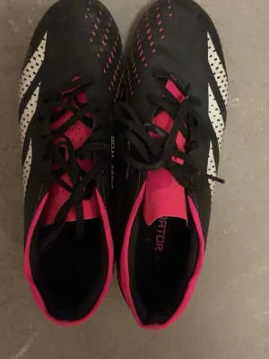 Adidas Predator svart/vit/rosa - Säljer ett par Adidas Predator fotbollsskor i svart med vita ränder och rosa detaljer. Skorna har snörning, mönstrad ovansida och dobbar för bra grepp på planen. Perfekta för dig som vill sticka ut med både stil och funktion på fotbollsplanen.