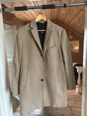 Beige ullkappa från Lindeberg - Stilren beige kappa från J.Lindeberg i ullblandning. Klassisk rak modell med slag, tre ytterfickor och knäppning framtill. Svart innerfoder ger en snygg kontrast. Perfekt för dig som gillar en clean och tidlös look.OBS: mitten knappen saknas på framsidan. 