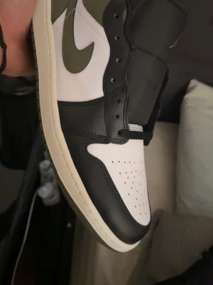 Nike Air Jordan 1 svart och olivgrön - Snygga Nike Air Jordan 1 sneakers i svart, vitt och olivgrönt läder. Klassisk high-top siluett med perforerad tåbox och ikonisk Swoosh-logga på sidan. Perfekta för dig som gillar streetwear och vill sticka ut med en tidlös modell. Dom är helt oanvända direkt från kartong 