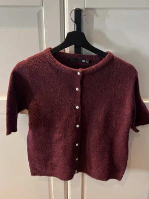Vinröd kofta med pärlknappar 157 - Vinröd stickad kofta från 157 med kort ärm och pärlknappar framtill. Koftan har rund halsringning och är gjord i ett mjukt, luddigt material. Perfekt för lager-på-lager och en trendig look.